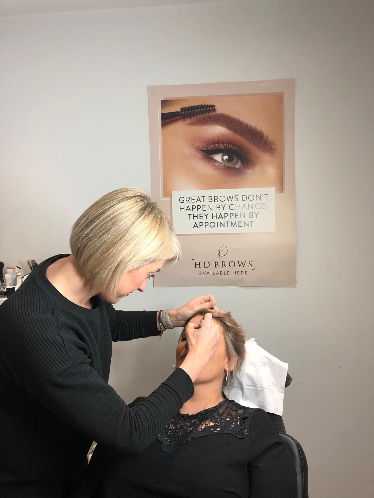 Nicola shaping a client’s brows beside an HD Brows poster
